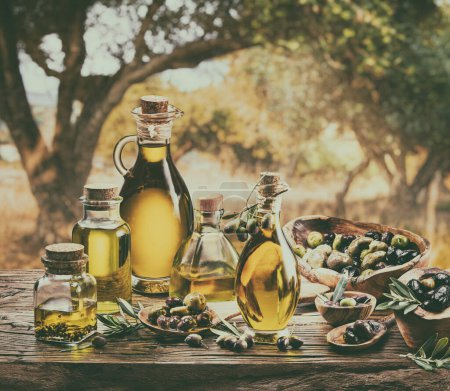 Foto de Botella de aceite de oliva de granja artesanal se encuentra en una mesa de madera, detrás hay un jardín de olivos en un día ligeramente bokeh, soleado. Imagen con estilo vintage. - Imagen libre de derechos