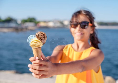 Photo for Happy, smiling girl holding ice cream cone with colorful ice cream balls. Sunny sea coastline at the background. - Royalty Free Image