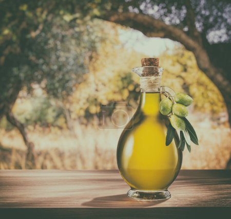 Téléchargez les photos : Une bouteille d'huile d'olive artisanale se trouve sur une table en bois, derrière un jardin d'oliviers par une journée légèrement bokeh et ensoleillée. Image avec style vintage. - en image libre de droit