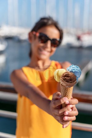 Téléchargez les photos : Fille heureuse et souriante tenant un cône de crème glacée avec des boules de crème glacée colorées. Côtes de mer ensoleillées en arrière-plan. - en image libre de droit
