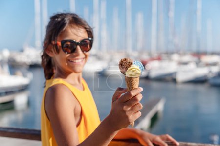 Téléchargez les photos : Fille heureuse et souriante tenant un cône de crème glacée avec des boules de crème glacée colorées. Côtes de mer ensoleillées en arrière-plan. - en image libre de droit
