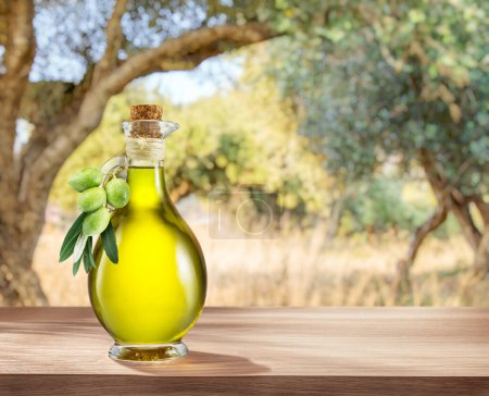 Foto de Botella de aceite de oliva de granja artesanal se encuentra en una mesa de madera, detrás hay un jardín de olivos en un día ligeramente bokeh, soleado. - Imagen libre de derechos
