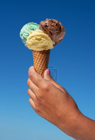 Foto de Cono de helado con bolas de helado de colores en la mano en el fondo del cielo azul. - Imagen libre de derechos