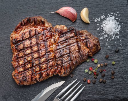 Photo for Grilled  delicious ribeye steak and some seasonings on black slate serving plate. Flat lay. - Royalty Free Image
