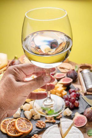 Téléchargez les photos : Un verre de vin blanc dans la main d'un homme avec une variété de fromages tranchés avec des fruits, de la menthe et des noix. Fond jaune. Magnifique fond de vin pour vos projets. - en image libre de droit