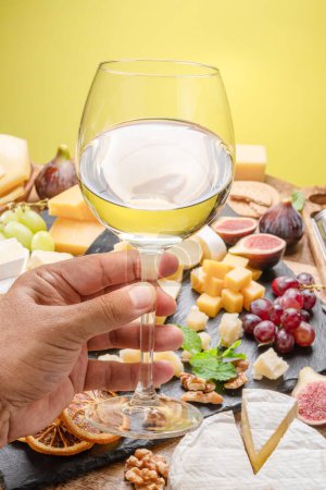 Photo for Glass of white wine in a man's hand with variety of sliced cheeses with fruits, mint and nuts. Yellow background. Wonderful wine background for your projects. - Royalty Free Image