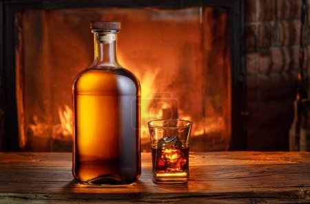 Photo for Glass of whiskey and whiskey bottle on old wooden table and blurred fireplace at the background. - Royalty Free Image