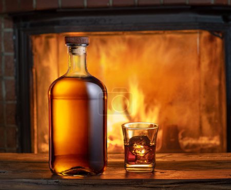 Foto de Vaso de whisky y botella de whisky en la vieja mesa de madera y chimenea borrosa en el fondo. - Imagen libre de derechos