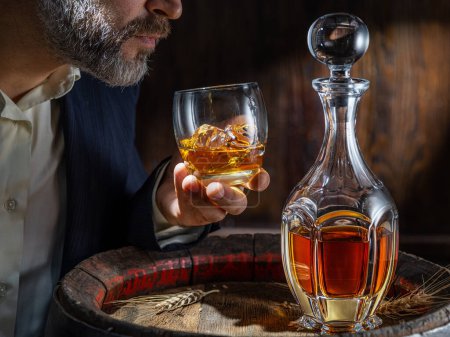 Téléchargez les photos : Dégustation de whisky. L'homme est assis devant un tonneau avec une carafe et un verre de whisky. - en image libre de droit