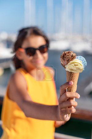 Photo for Happy, smiling girl holding ice cream cone with colorful ice cream balls. Sunny sea coastline at the background. - Royalty Free Image