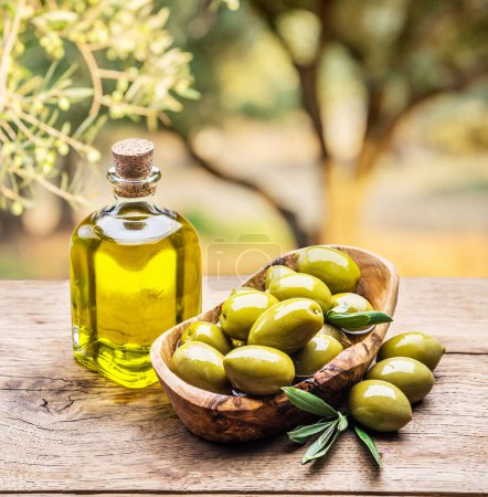 Photo for Olive oil in a bottle with a bowl of green olives on a wooden vintage table against the backdrop of an olive garden on a sunny summer day. - Royalty Free Image