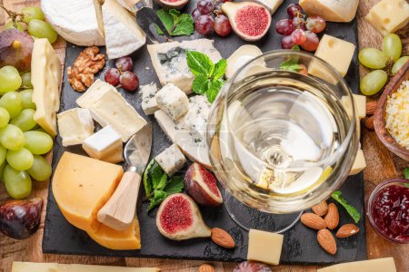 Photo for Glass of white wine with variety of sliced cheeses with fruits, mint, nuts and cheese cutting knives. Wonderful wine and cheese background for your projects. - Royalty Free Image