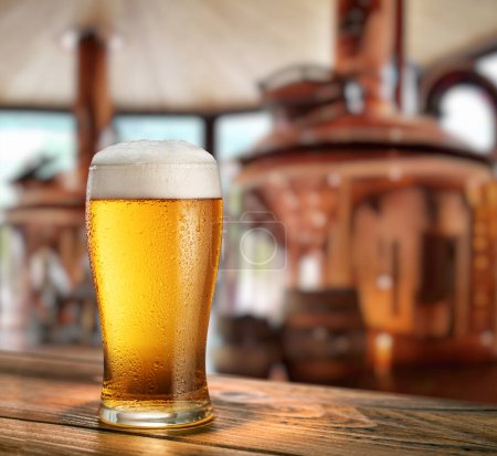 Foto de Vidrio refrigerado de cerveza ligera sobre mesa de madera y sistema de elaboración de cerveza de cobre difuminado en el fondo. - Imagen libre de derechos