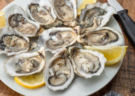Opened raw oysters with sauce and lemon slices on plate on wooden table. Top view.