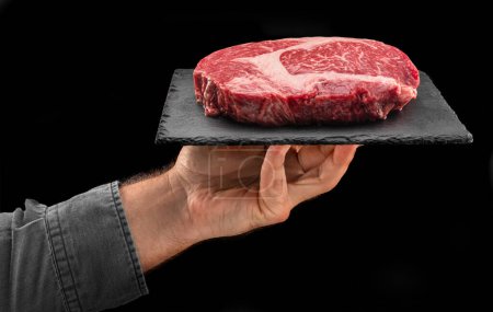 Téléchargez les photos : Cook tient une plaque de service en graphite avec un steak de côtelette cru sur fond noir. - en image libre de droit