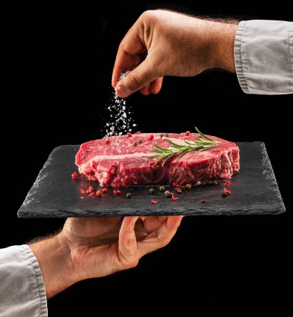 Photo for Chef is salting or seasoning raw ribeye steak laying on graphite serving board. Black background. - Royalty Free Image