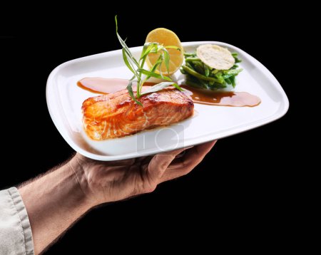 Foto de Chef sostiene plato blanco de salmón asado con guarnición de judías verdes sobre fondo negro. Sabroso fondo de la comida. - Imagen libre de derechos