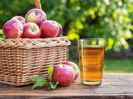 Foto de Vaso de zumo de manzana fresca o sidra y cesta con manzanas maduras sobre una mesa de madera en el huerto. - Imagen libre de derechos
