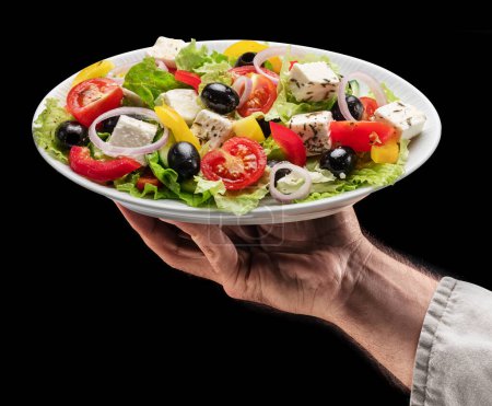 Foto de Cook sostiene un plato blanco de ensalada griega sobre fondo negro. Sabroso fondo de la comida. - Imagen libre de derechos