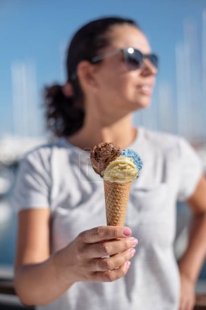 Foto de Chica feliz y sonriente sosteniendo cono de helado con bolas de helado de colores. Línea de costa soleada al fondo. - Imagen libre de derechos
