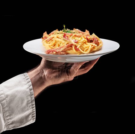 Foto de Cook sostiene un plato blanco de espaguetis con tocino o pasta carbonara sobre fondo negro. Sabroso fondo de la comida. - Imagen libre de derechos