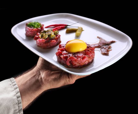 Photo for Chef holds white plate of steak tartare on black background. Tasty food background. - Royalty Free Image