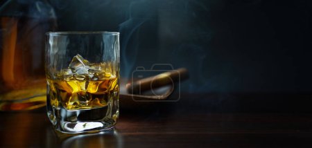 Foto de Un vaso de whisky con hielo sobre una mesa de madera, un cigarro cubano humeante y una botella de whisky en un ambiente ahumado de un bar nocturno - Imagen libre de derechos