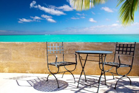 Foto de An empty street bar, cafe or restaurant with an old wrought iron table and chairs standing near a colonial stone wall overlooking a seascape with palm trees. Tropics, summer, sunny day - Imagen libre de derechos
