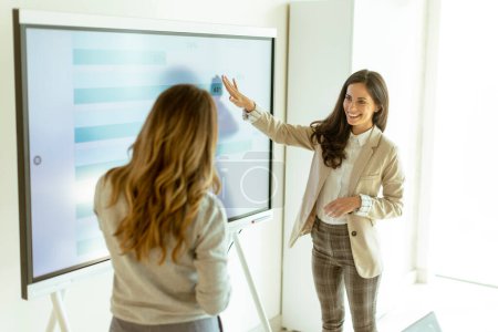 Photo for Two young business women discussing financial results on the big wall screen in the office - Royalty Free Image
