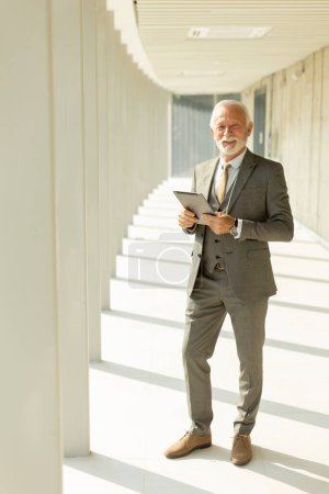 Téléchargez les photos : Bel homme d'affaires mature debout avec tablette numérique dans le couloir de bureau - en image libre de droit