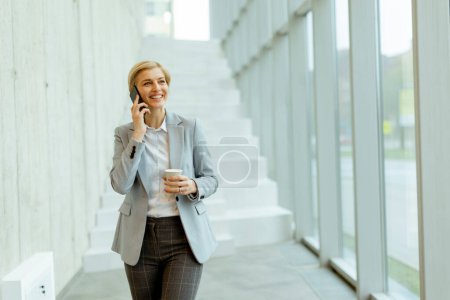 Foto de Mujer de negocios usando el teléfono móvil mientras camina en el pasillo de la oficina moderna - Imagen libre de derechos