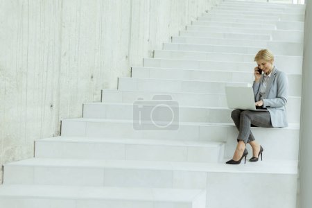 Photo for Businesswoman using mobile phone while working on laptop on the modern office stairs - Royalty Free Image