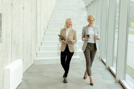 Téléchargez les photos : Deux femmes d'affaires marchant avec tablette numérique et téléphone portable dans le couloir du bureau - en image libre de droit