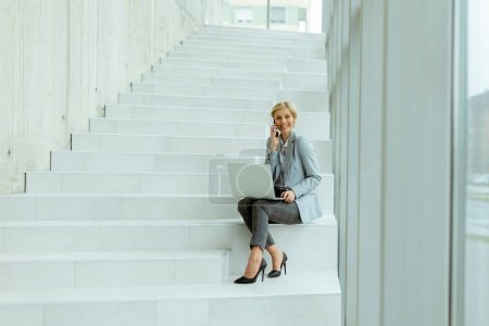 Photo for Businesswoman using mobile phone while working on laptop on the modern office stairs - Royalty Free Image