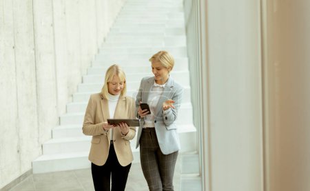 Téléchargez les photos : Deux femmes d'affaires marchant avec un ordinateur portable et un téléphone portable dans le couloir du bureau - en image libre de droit