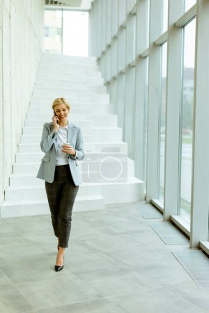 Téléchargez les photos : Femme d'affaires utilisant un téléphone portable tout en marchant sur le couloir de bureau moderne - en image libre de droit