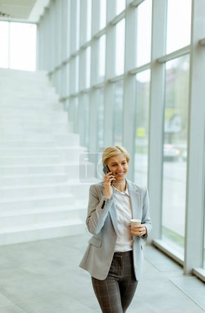 Téléchargez les photos : Femme d'affaires utilisant un téléphone portable tout en marchant sur le couloir de bureau moderne - en image libre de droit