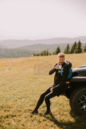 Photo for Handsome young man relaxing and drinking coffee by the terrain vehicle hood at countryside - Royalty Free Image