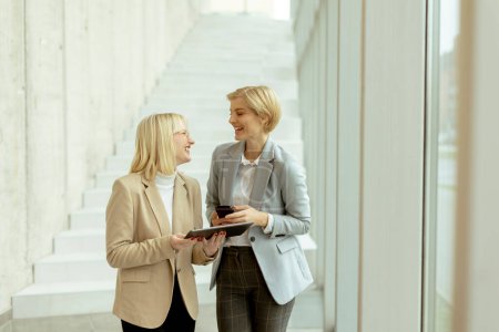 Foto de Dos mujeres de negocios caminando con portátil y teléfono móvil en el pasillo de la oficina - Imagen libre de derechos