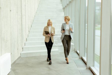 Photo for Two business women walking with digital tablet and mobile phone in the office corridor - Royalty Free Image