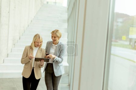 Foto de Dos mujeres de negocios caminando con portátil y teléfono móvil en el pasillo de la oficina - Imagen libre de derechos