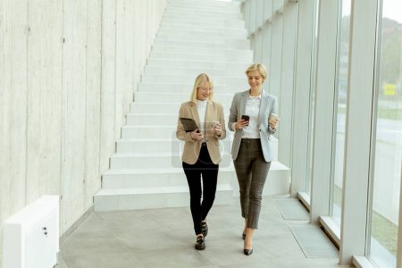 Foto de Dos mujeres de negocios caminando con tableta digital y teléfono móvil en el pasillo de la oficina - Imagen libre de derechos