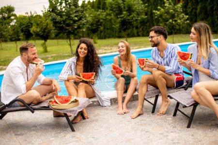 Téléchargez les photos : Group of young people sitting by the swimming pool and eating watermelon in the house backyard - en image libre de droit