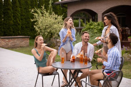 Foto de Group of happy young people cheering with cider and eating pizza by the pool in the garden - Imagen libre de derechos