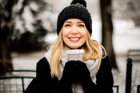Téléchargez les photos : Jolie jeune femme en vêtements chauds jouissant dans la neige avec tasse à café à emporter - en image libre de droit