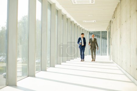 Foto de Young y un hombre de negocios mayor caminan por el pasillo de una oficina, en lo profundo de la conversación. Ambos están vestidos profesionalmente, reflejando su perspicacia y estatus empresarial. Están navegando por las complejidades del mundo corporativo, encontrando soluciones y - Imagen libre de derechos