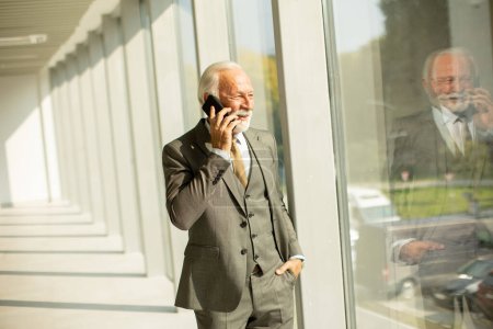 Téléchargez les photos : Un homme d'affaires senior se tient dans un couloir de bureau, concentré sur son téléphone portable. Il est vêtu de vêtements formels, exsudant confiance et professionnalisme - en image libre de droit