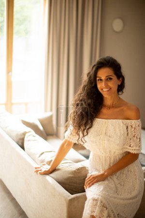 Téléchargez les photos : Pretty young woman with curly hair is relaxing in the comfort of her own room, dressed in a white flowing dress - en image libre de droit