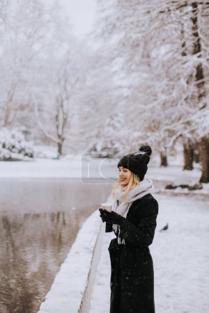 Téléchargez les photos : Pretty young woman in warm clothes enjoying in snow with takeaway coffee cup - en image libre de droit