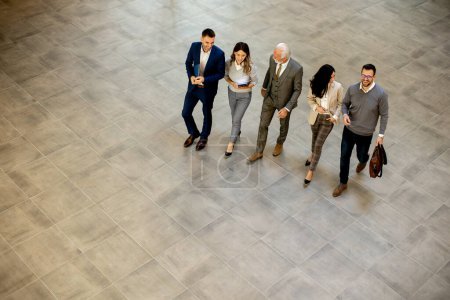 Foto de Group of young and senior business people are walking in an office hallway, captured in an aerial view. They are dressed in formal attire, walking with purpose and intent and discussing amongst themselves and interacting with colleagues - Imagen libre de derechos